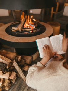 Lectrice assise dans un fauteuil, livre ouvert dans les mains, à ses pieds du bois coupé pour le poêle devant elle où un feu crépite